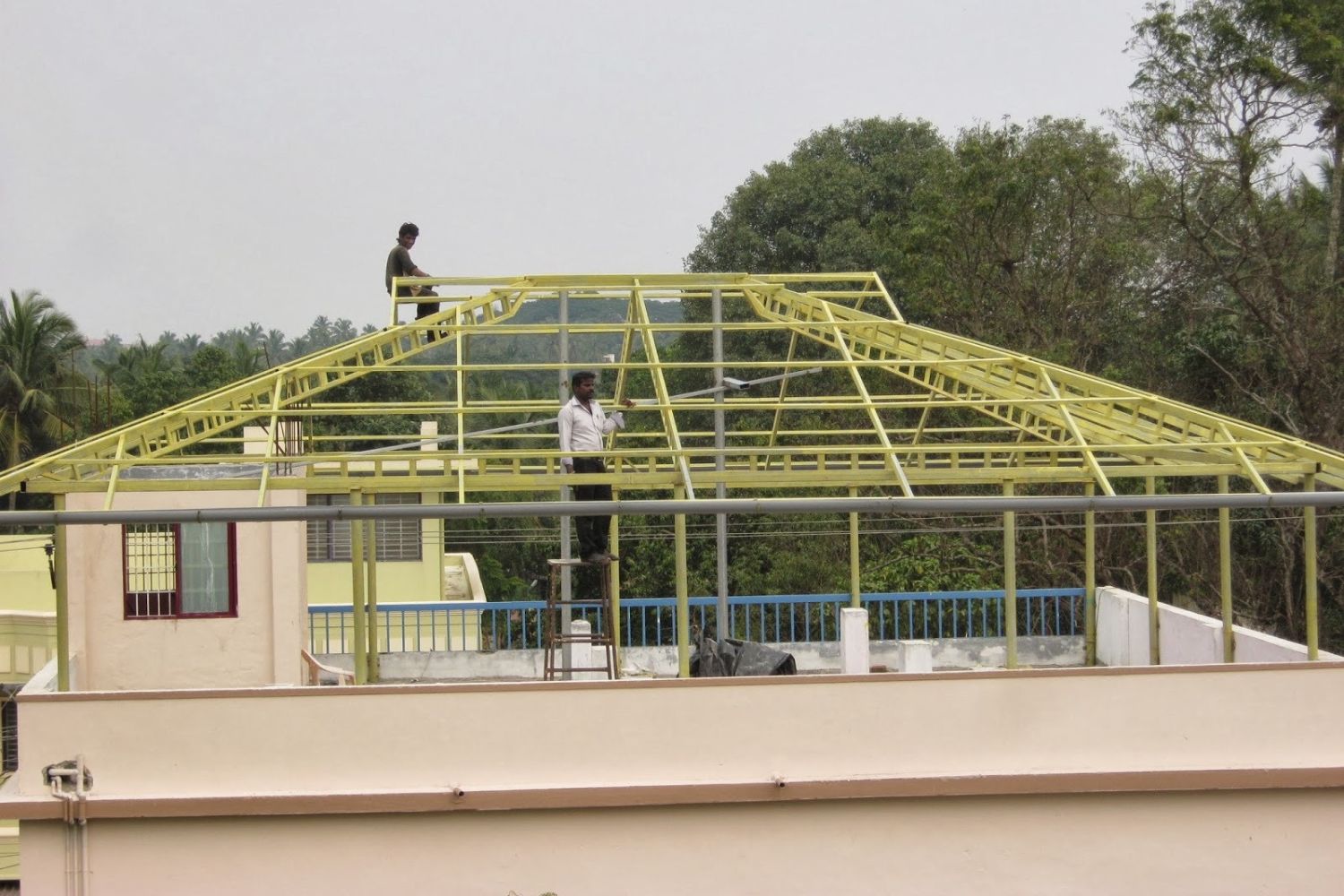 Two workers assembling a yellow steel roof truss frame on top of an unfinished concrete rooftop, hipped truss profile against trees and sky