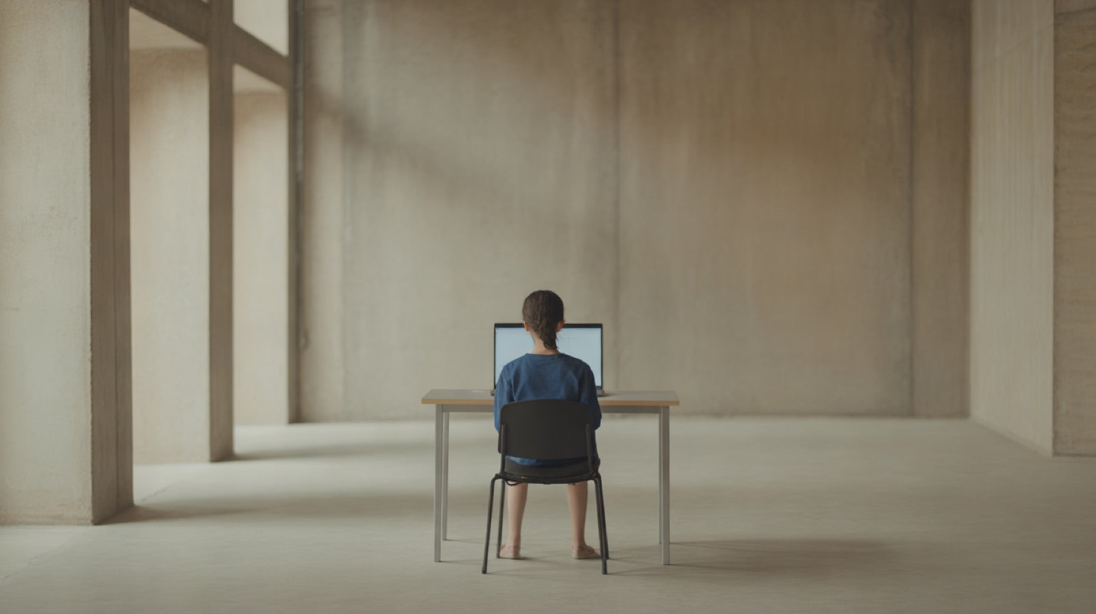 AutoCAD for Beginners: a student seen from behind sitting at a plain table with an open laptop whose screen is a plain bright glow, bare wall
