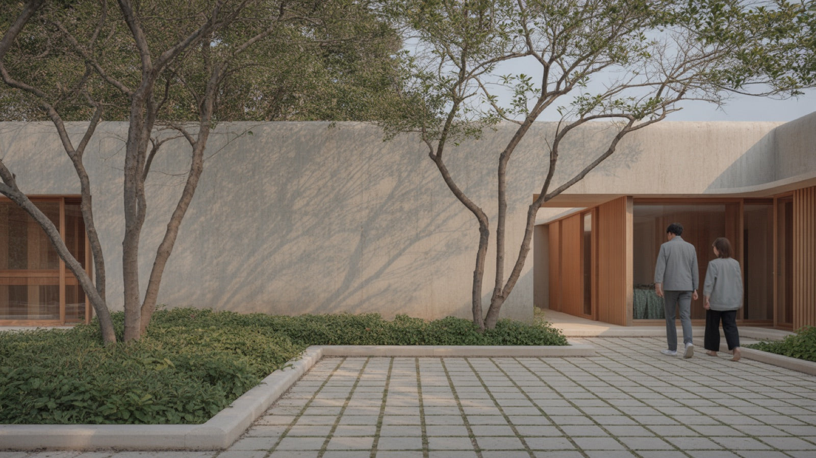Low concrete and timber building beside a planted courtyard with trees and stone paving, two people walking the path