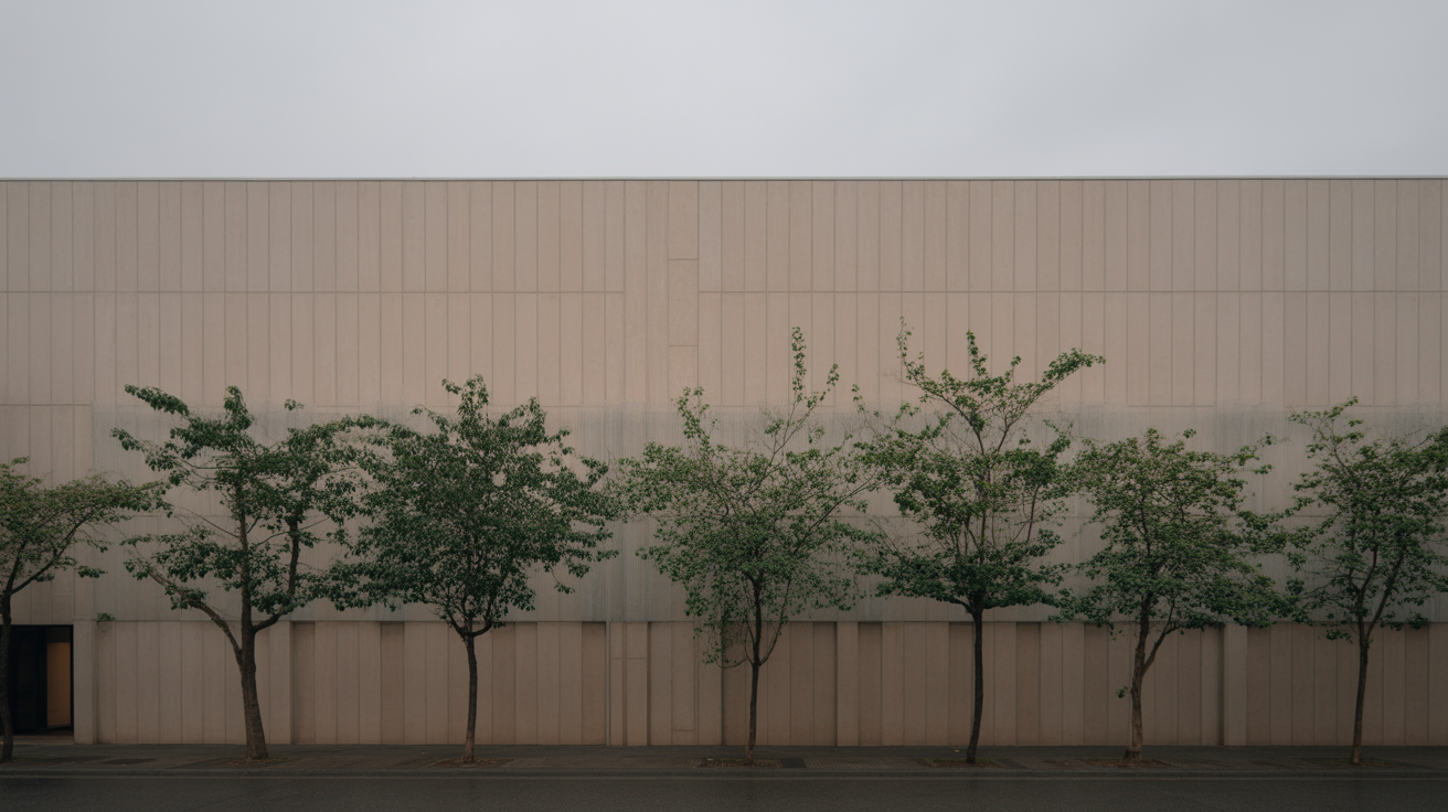 A row of street trees along a plain building facade in flat overcast light, foliage silhouettes clear